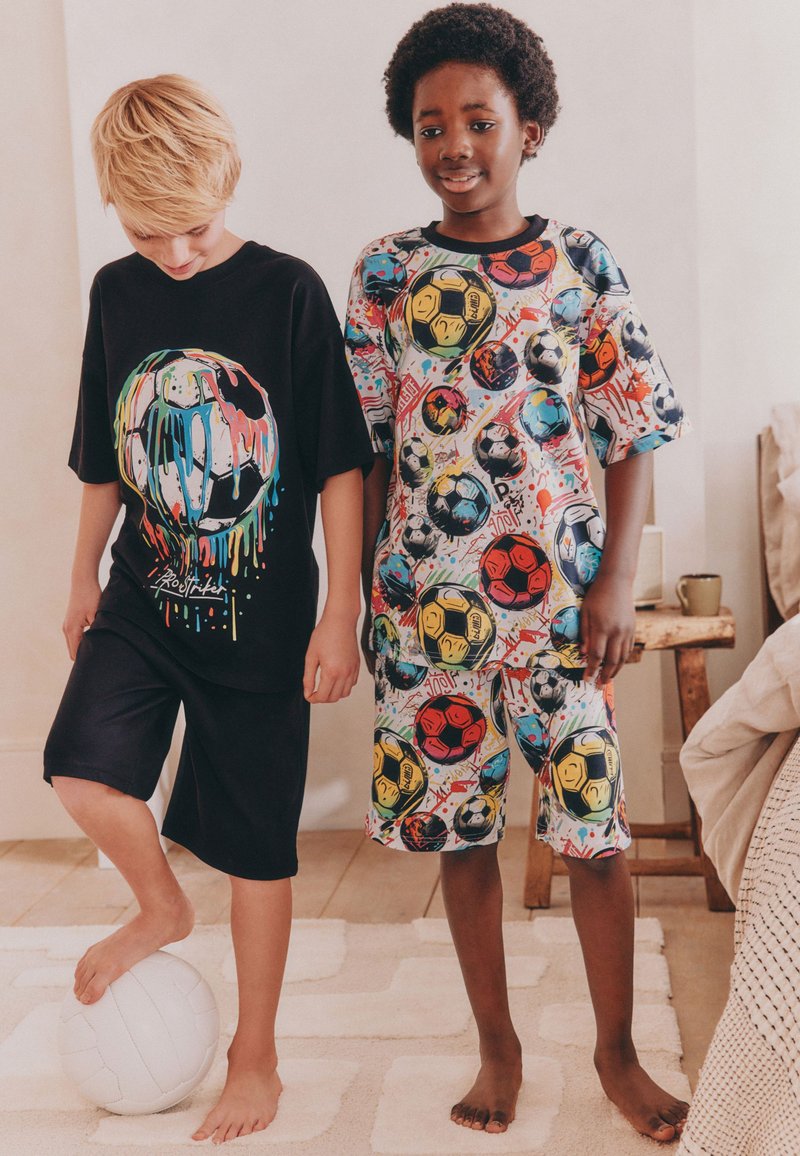 To drenge i farverige fodboldtema outfits står barfodet indendørs, hvor den ene balancerer en hvid fodbold med sin fod på et lyst tæppe.