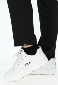 Witte platform sneakers met vetersluiting, textuur met een profiel, Fila-logo en contrasterende rode en blauwe accenten. Zwarte broek zichtbaar.