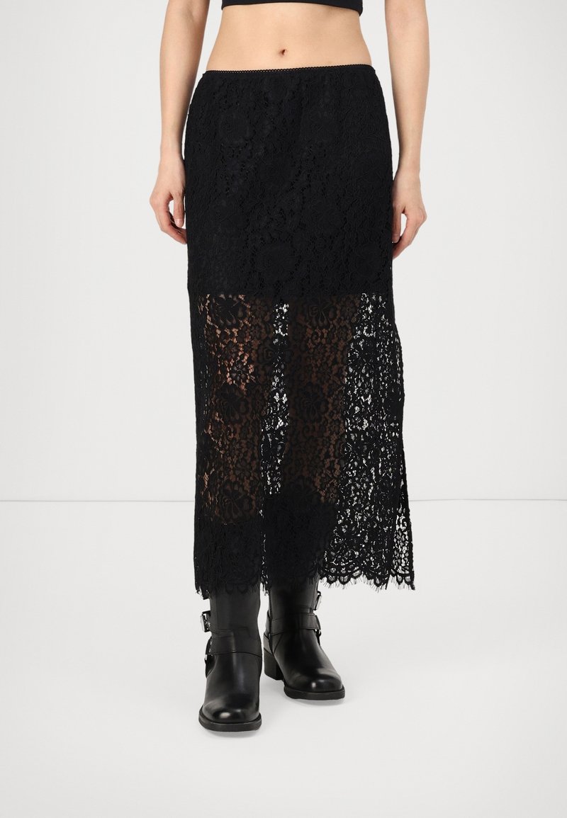 Jupe midi en dentelle noire avec un motif floral transparent à partir du milieu de la cuisse, portée avec des bottines noires.