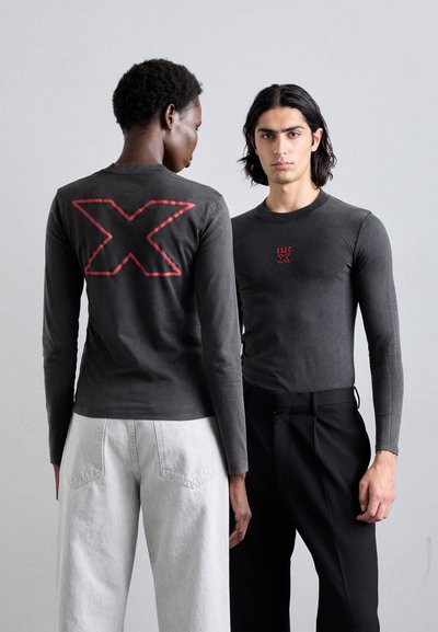 Langärmelige graue Hemden haben ein rotes gesticktes "X" auf dem Rücken und ein kleines Logo auf der Vorderseite; das Modell trägt schwarze Hosen und hellgraue Jeans.