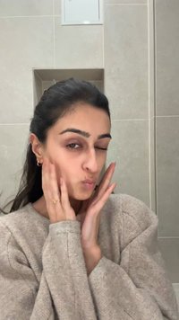 Une personne portant un pull en tricot beige, aux cheveux foncés, faisant un clin d'œil et touchant son visage, devant un fond de carreaux gris clair.