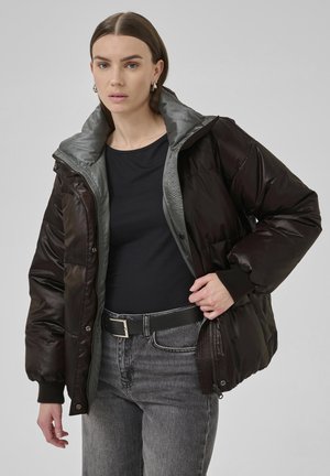 Eine Frau passt eine dunkelbraune Daunenjacke mit einem grauen Innenfutter an. Sie trägt ein schwarzes Oberteil, graue Jeans und einen schwarzen Gürtel und steht vor einem grauen Hintergrund.