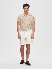 Shorts en lin beige clair avec taille élastique ; chemise à manches courtes en crème avec imprimé de feuilles beiges ; chaussures marron foncé avec détails texturés.