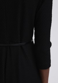 Vestido negro con textura y silueta ajustada, con mangas tres cuartos y un cinturón negro delgado en la cintura.