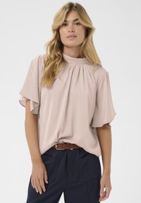 Blouse rose pâle à manches courtes avec un col rond et des détails froncés, associée à un pantalon bleu foncé et une ceinture marron. Texture lisse.