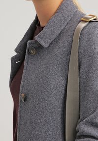 Kvinna iklädd en grå ullkappa över en mörk tröja, bär en beige väska med ett guldspänne på axeln.