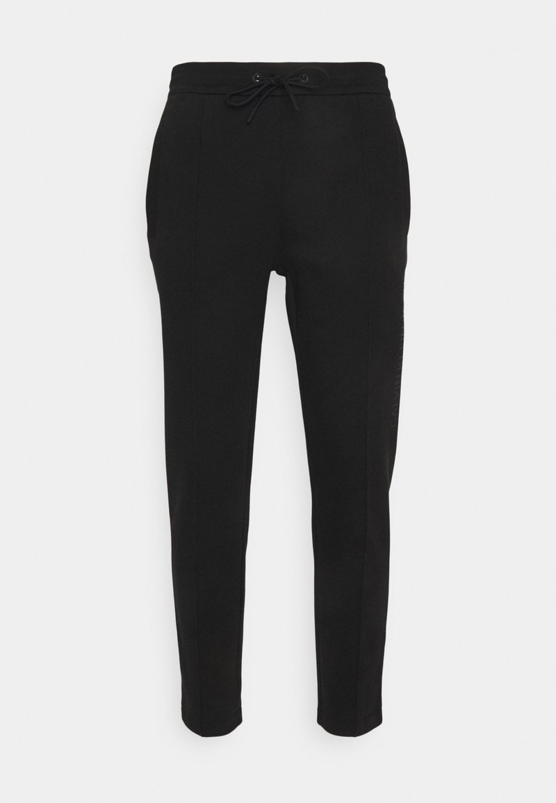 Černé tepláky mají stahovací pas, zúžené nohavice, boční kapsy a diskrétní detail loga na levé straně. Hladká textura.