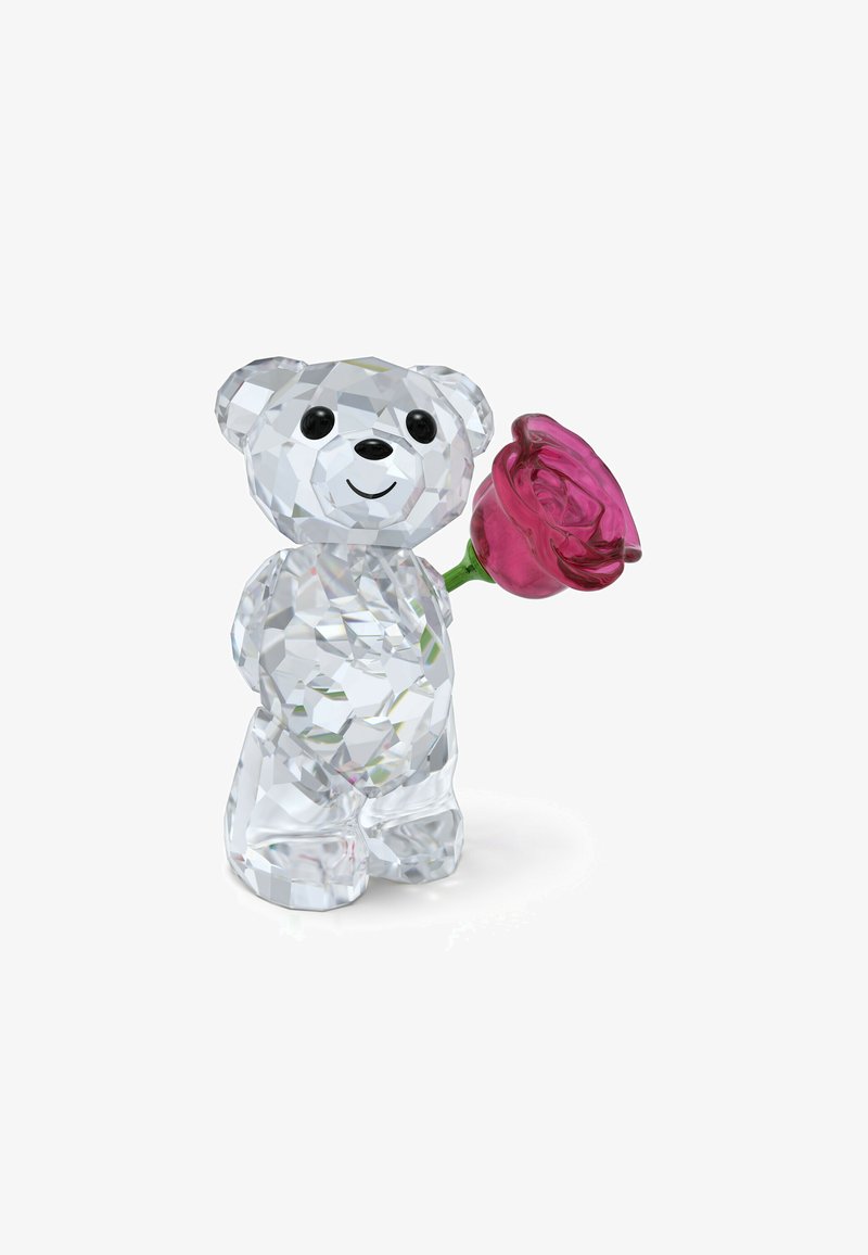 Figurka niedźwiedzia wykonana z kryształu, z oszlifowaną powierzchnią, czarnymi oczami i różą w kolorze różowym w prawej łapie. Przezroczysty i lustrzany materiał, kompaktowy kształt.