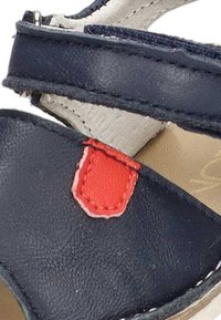 Sandalia de piel azul marino con un acento rojo, correa ajustable y suela beige. Textura suave con detalles de costura.