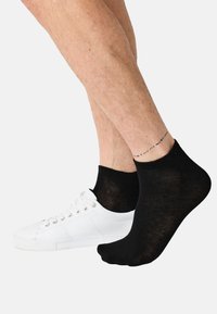 Sort ankelstrømpe på en fod ved siden af en hvid sneaker, der fremhæver en tynd stoftekstur og subtil glans. Hudtonen er lys med fint hår.