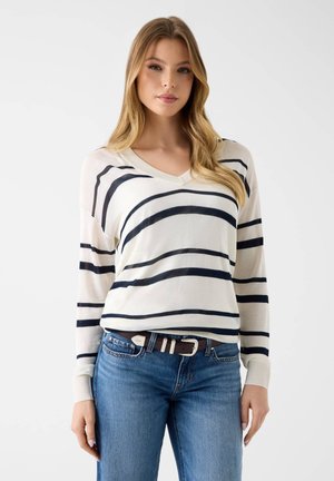 Mujer con suéter blanco de cuello en V con rayas horizontales negras y vaqueros azules, de pie frente a un fondo claro y liso.