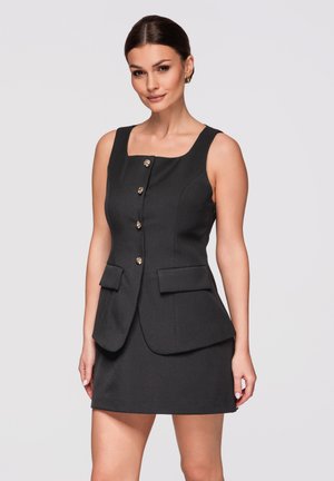 Kobieta w czarnej, bezrękawnikowej sukience z kwadratowym dekoltem, złotymi guzikami z przodu oraz dwoma przednimi kieszeniami z klapkami, stojąca na jednolitym tle.