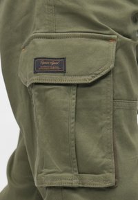 Χακί παντελόνια με μεγάλη πλαϊνή τσέπη και υφασμάτινη ετικέτα με σκουρό μπλε φόντο και πορτοκαλί κείμενο. Λεπτή υφή και ανθεκτική κατασκευή.