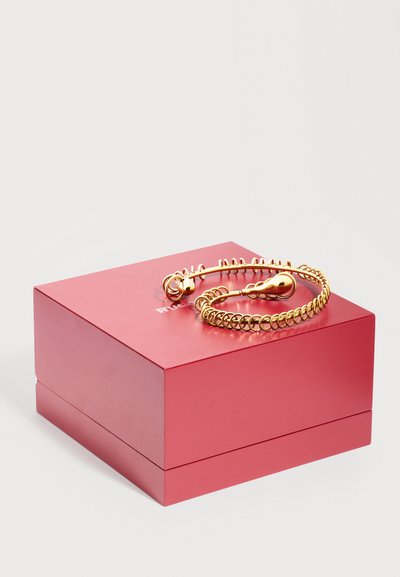 Βραχιόλι σε χρυσό χρώμα, τυλιγμένο με σφαιρικά άκρα, τοποθετημένο πάνω σε ένα κλειστό ματ κόκκινο τετράγωνο κουτί κοσμημάτων, με φόντο απλό λευκό.