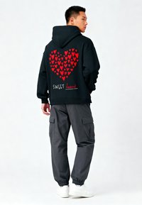 Felpa nera con cappuccio e motivo a cuori rosse, testo "SWEET heart". Indossata con pantaloni cargo grigi e sneakers bianche. Design semplice, vestibilità casual.