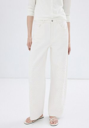 Femme portant un jean taille haute à jambes larges blanc avec broderies florales, associé à des sandales plates blanches à lanières et un haut blanc boutonné.