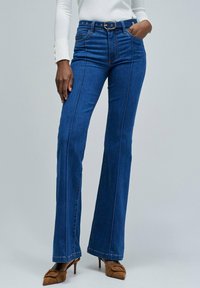 Femme portant un jean bleu évasé taille haute avec des coutures frontales, un haut blanc à manches longues, des mules à talons bruns et une ceinture noire avec boucle dorée.