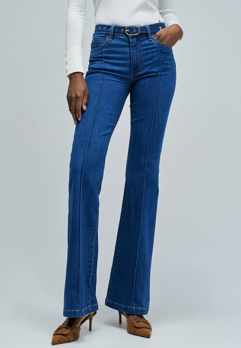 Femme portant un jean bleu évasé taille haute avec des coutures frontales, un haut blanc à manches longues, des mules à talons bruns et une ceinture noire avec boucle dorée.