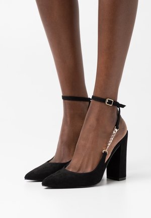 Escarpins noirs en daim à talon haut avec un bout pointu, une sangle à la cheville et un accent en chaîne dorée sur le côté ; le talon carré offre de la stabilité.