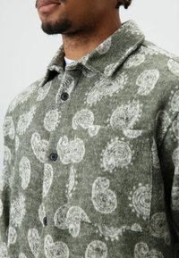 Grünes Wollhemd mit weißem Paisley-Muster, einer Fronttasche, schwarzen Knöpfen, strukturiertem Kragen und weicher Textur.