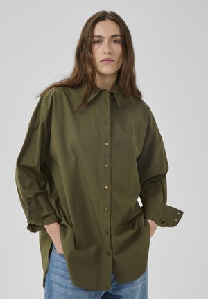 Ženska z dolgimi rjavimi lasmi, oblečena v preveliko kaki zeleno srajco z gumbi in modre kavbojke, roke v žepih, nevtralen izraz, sivo ozadje.