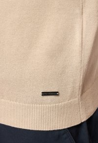 Maglione beige lavorato a maglia con orlo a coste e pannelli laterali. Presenta un'etichetta in metallo nero vicino al fondo. Tessuto liscio con un design leggero.
