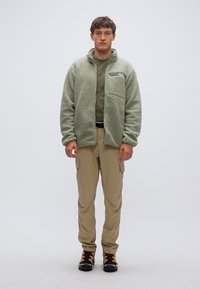 Φλις μπουφάν σε απαλό πράσινο, με φερμουάρ στο μπροστινό μέρος, τσέπη στο στήθος, σε συνδυασμό με μπεζ παντελόνι cargo και μαύρα/κόκκινα παπούτσια πεζοπορίας. Απλό φόντο.