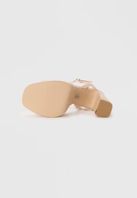 Beige korkokenkä, jossa on teksturoitu pohja, leveä hihna ja nilkkasolki. Kannan korkeus on noin 8 cm ja se on sileäpintainen.