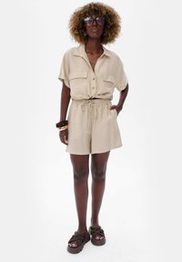 Beige short-sleeve button-up shirt met twee zakken, gecombineerd met bijpassende shorts. Draag het met bruine sandalen en gestapelde armbanden.