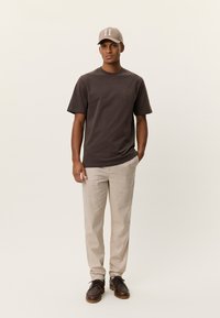 Καφέ κοντομάνικη μπλούζα με τσέπη στο στήθος, εκρού παντελόνι και μπεζ καπέλο με ανοιχτό γκρι αριθμό "II" στο μπροστινό μέρος. Καφέ παπούτσια.