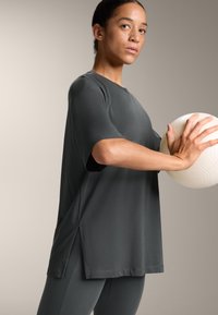 Top sportiv scurt cu mânecă scurtă, de culoare gri închis, cu fante pe laterale, purtat cu colanți texturați asortați. Modelul ține în mână o minge cu un model.