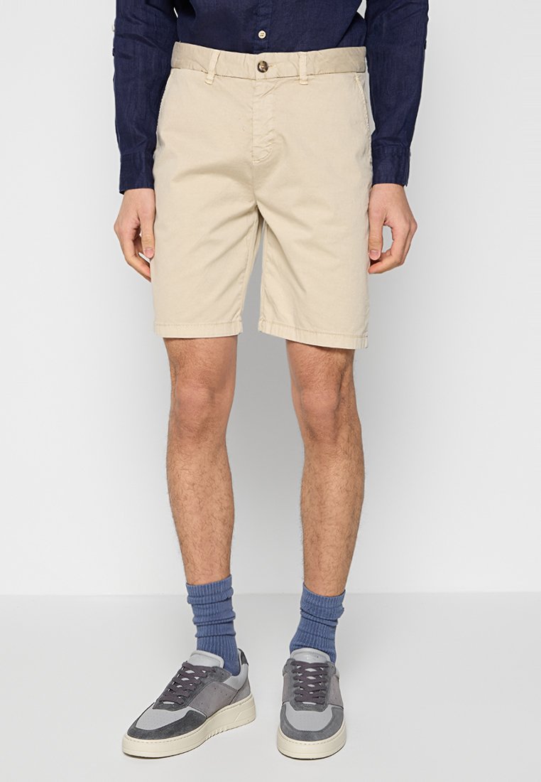 Beżowe bawełniane szorty z zapinanym paskiem, kieszeniami po bokach i podwiniętym dołem. Połączone z szarymi trampkami i niebieskimi skarpetami w prążki.