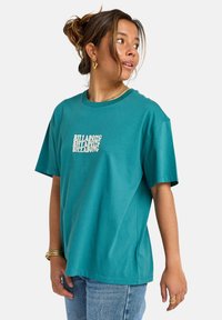 Turquoise cotton t-shirt with a relaxed fit, crew neck, short sleeves, and a repeating white "BILLABONG" logo on the front.