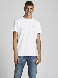 Camiseta de algodón blanca de cuello redondo con mangas cortas, que presenta un pequeño logo negro en el lado izquierdo del pecho. Combinada con unos vaqueros azules. Diseño simple.