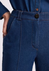 Zbliżenie na osobę ubraną w niebieskie dżinsy z pomarańczowymi przeszyciami, jedna ręka spoczywa w przedniej kieszeni.