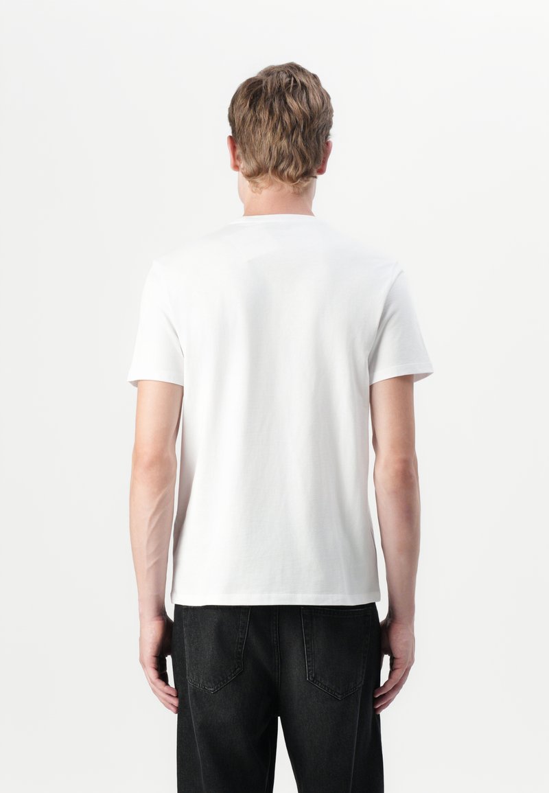 T-shirt blanc à manches courtes, col rond, en coton, avec une texture lisse. Porté avec un jean en denim noir avec des poches arrière.