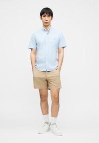 Homme portant une chemise à manches courtes bleu clair boutonnée, un short beige, des chaussettes blanches et des baskets blanches, debout les mains dans les poches.