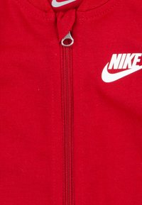 Piros, cipzáros ruházat sima anyagból, középső cipzárral és fehér Nike logóval. Rugalmas, testhezálló kialakítás kerek nyakkal.