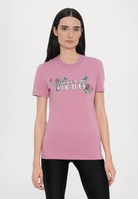 T-shirt di cotone rosa chiaro con maniche corte, caratterizzata dal logo "VERSACE JEANS COUTURE" e da disegni floreali con accenti blu e verde.