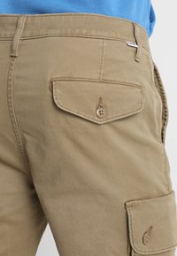 Khaki cargo shorts valmistettu sileästä puuvillakankaasta, jossa on koristeellinen läppätasku napilla ja tuplanomisteet.