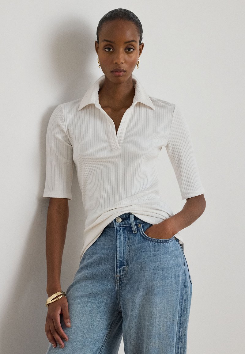 Γυναίκα που φοράει λευκό ριμπ από μετάξι πόλο με μισά μανίκια, ανοιχτό μπλε ψηλόμεσα τζιν, χρυσά σκουλαρίκια και χρυσό βραχιόλι, στέκεται με το χέρι στην τσέπη.