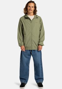 Lichtgroene, ritsjacket met een capuchon en elastische manchetten; gedragen over losse, blauwe jeans. Zwarte schoenen maken de outfit compleet.