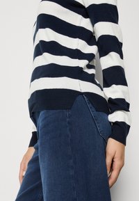 Personne portant un pull rayé bleu marine et blanc à manches longues et un jean bleu foncé, debout devant un fond clair.