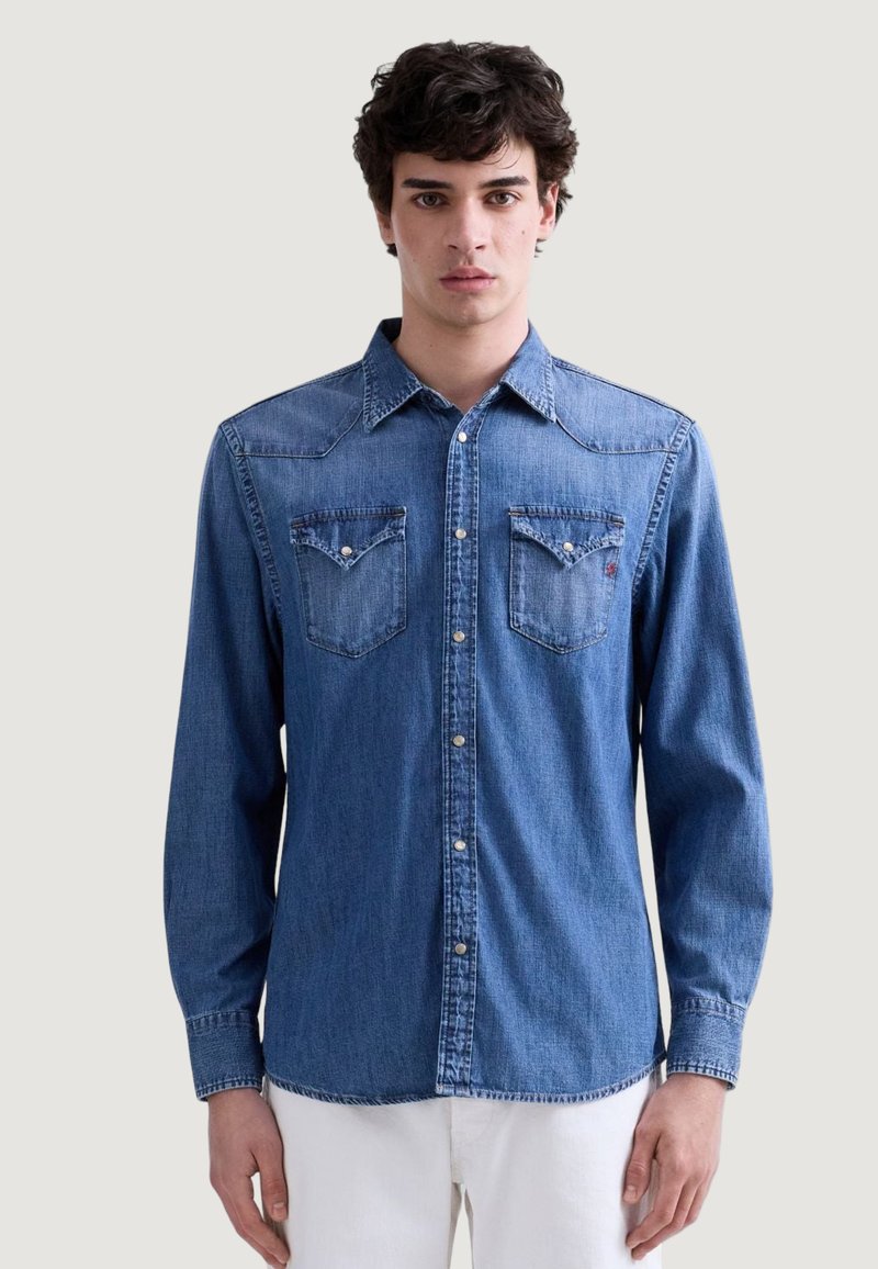 Jonge man die een gesloten blauwe denim blouse met parelmoer snapzakken draagt, staand tegen een effen lichte achtergrond.