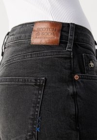Jeans in denim nero con un'etichetta in pelle marrone con la scritta "SCOTCH & SODA AMSTERDAM", arricchiti da cuciture decorative blu e accessori in rame.