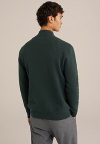 Tumši zaļš adīts džemperis ar augsto apkakli un ribotām piedurknēm, kas saskaņots ar gaiši pelēkiem biksēm. Audums izskatās maigs un teksturēts.