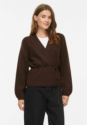 Femme portant un pull cache-cœur marron avec une ceinture nouée sur un haut blanc et un pantalon noir, debout devant un fond clair uni.