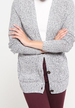 Personne portant un cardigan en maille gris avec de gros boutons par-dessus un haut blanc et un pantalon rouge foncé, les bras croisés sur la poitrine.