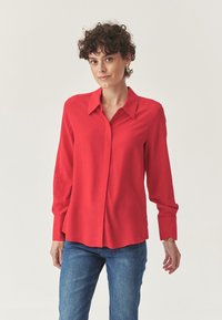Camisa roja de manga larga con cuello clásico, cierre de botones y tejido suave. Combinada con vaqueros azules, expuesta sobre un fondo claro.