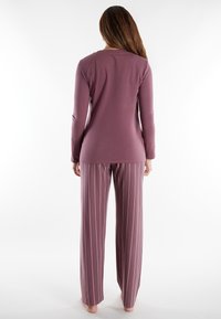 Woman standing barefoot wearing a long-sleeve mauve top and mauve striped wide-leg pants, viewed from the back against a white background.